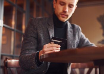 Portrait of modern young guy in formal clothes that sits in the cafe and holds credit card in hand.